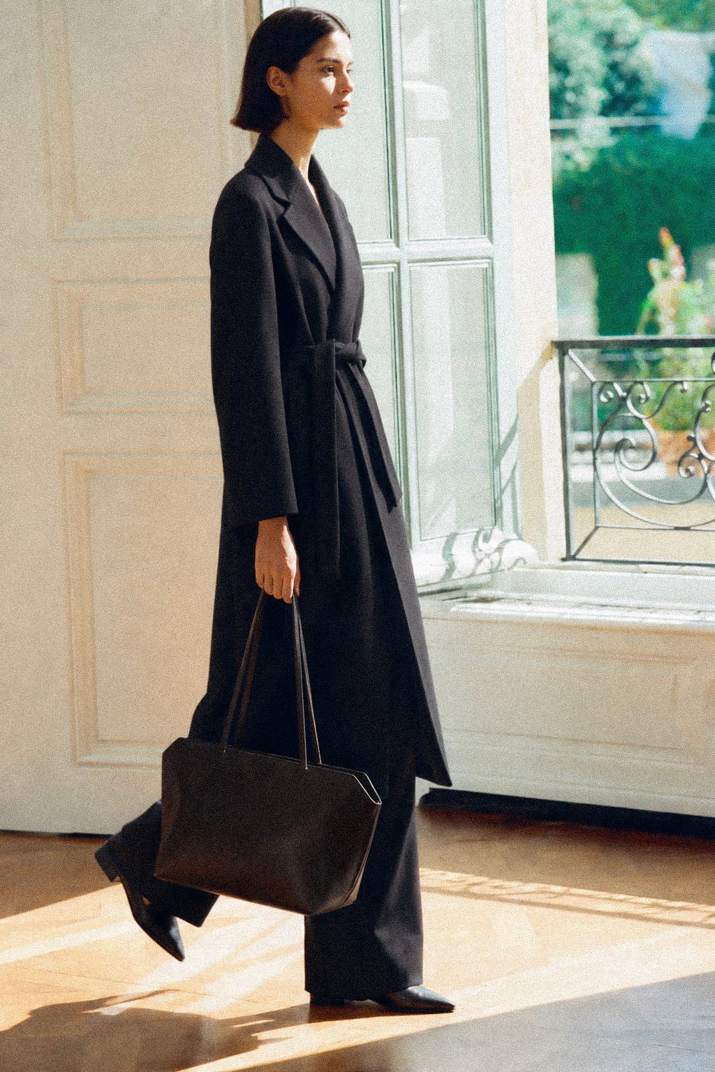 A woman in a long black belted coat and wide-leg trousers walks indoors while carrying a large black tote bag near a sunlit window.